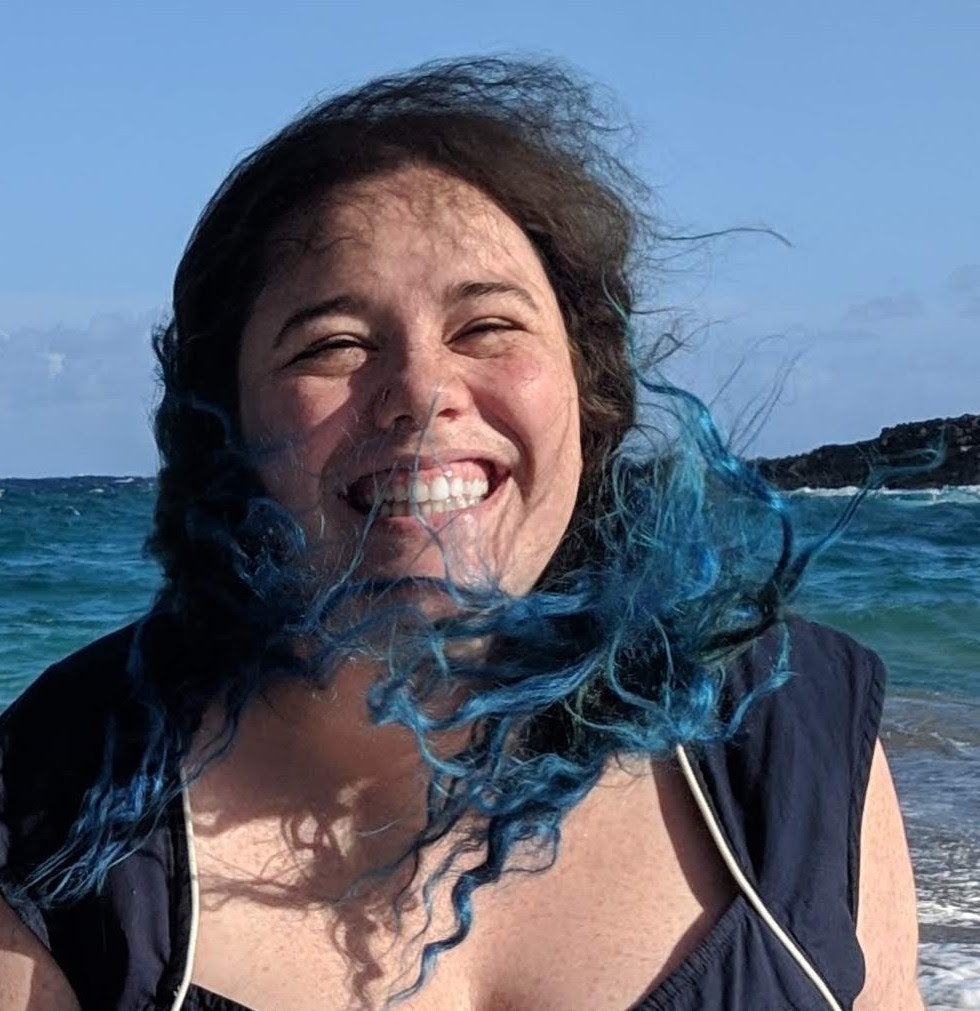 Tiffany, a white woman with curly blue and brown hair laughs in front of a blue ocean, and blue sky