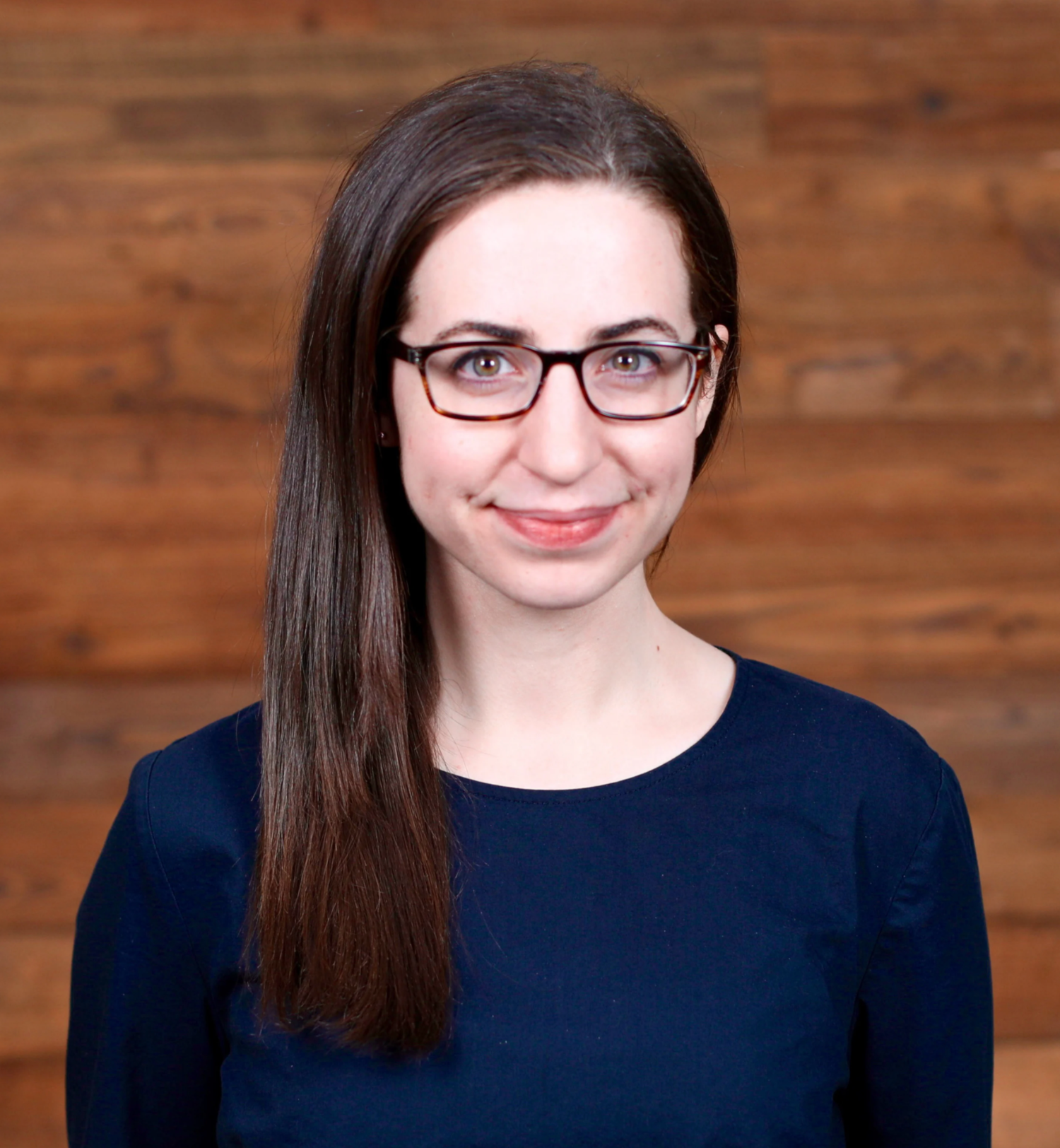 woman with long and straight brown hair, glasses, and a navy blue blouse smiling in front of a wooden background