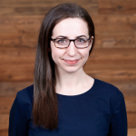 woman with long and straight brown hair, glasses, and a navy blue blouse smiling in front of a wooden background