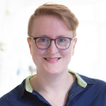 Ruth is wearing a dark blue shirt with a pale green trim inside the collar. She is smiling at the camera and is wearing glasses.